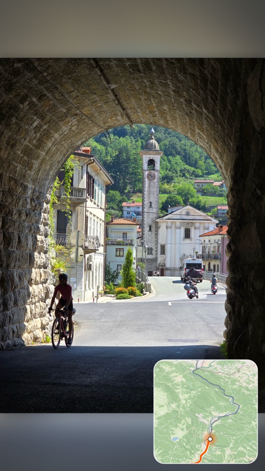 A film frame filled by a photograph of a rider in a stone tunnel, a church tower in the sunlit village beyond. In the bottom corner, a small map tile carries the orange route.